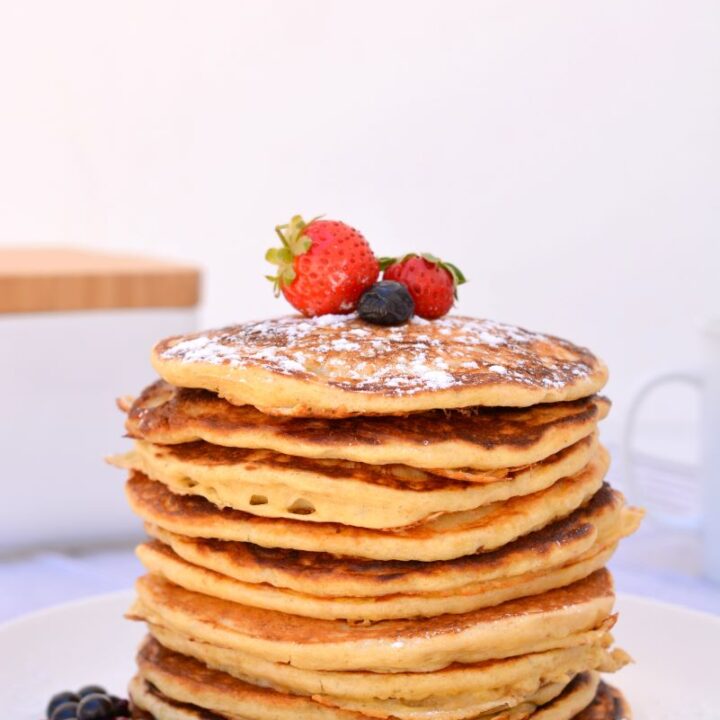 Stapel versgebakken American Pancakes met zuurdesem op een wit bord gedecoreerd met aardbeien en bosvruchten. Op de achtergrond staan een witte zoutpot met houten deksel en een wit kopje.
