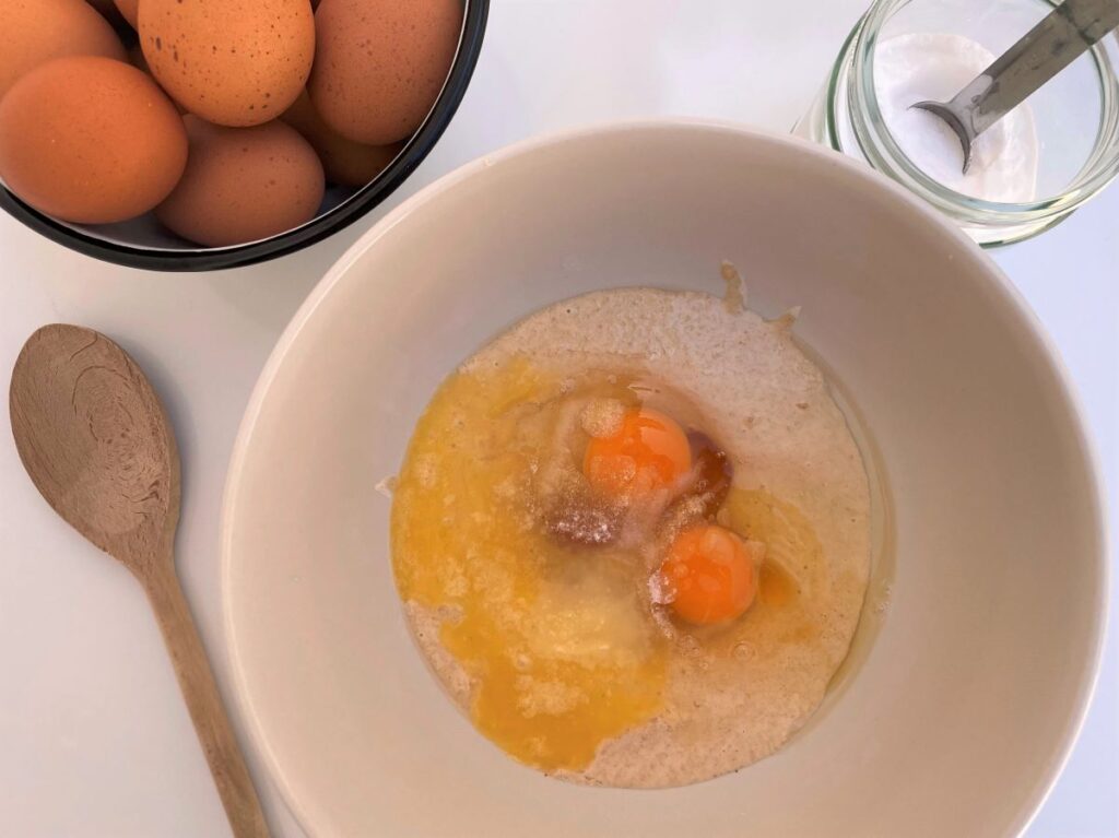 Zuurdesem pannenkoeken beslag in een witte kom naast een kom met eieren en een potje baking soda