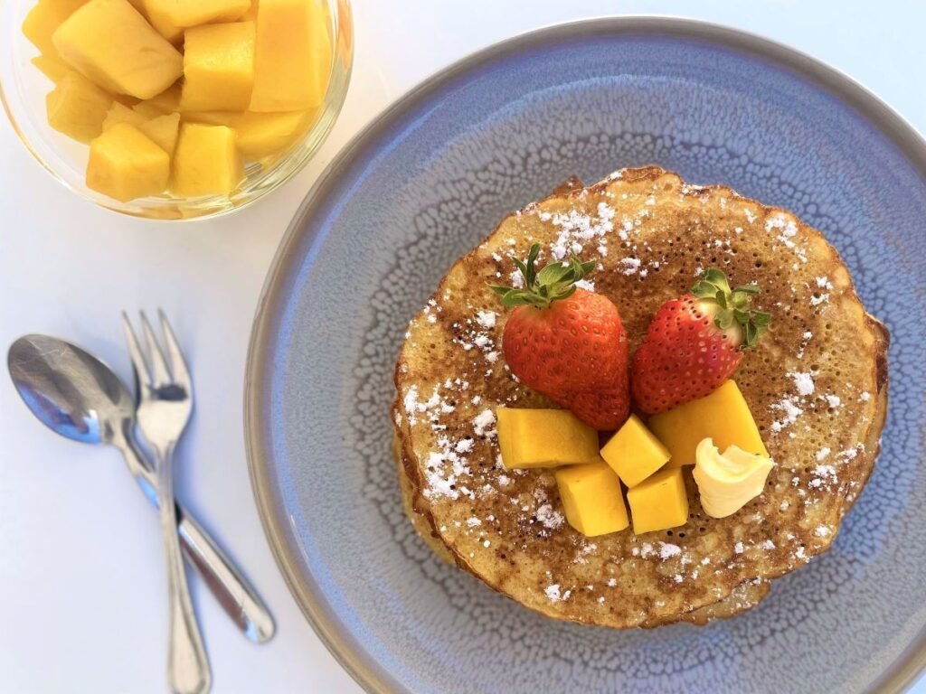 Zuurdesem pannenkoeken bedekt met aardbeien, mango en roomboter op een blauw bord, naast een glazen kommetje met mango