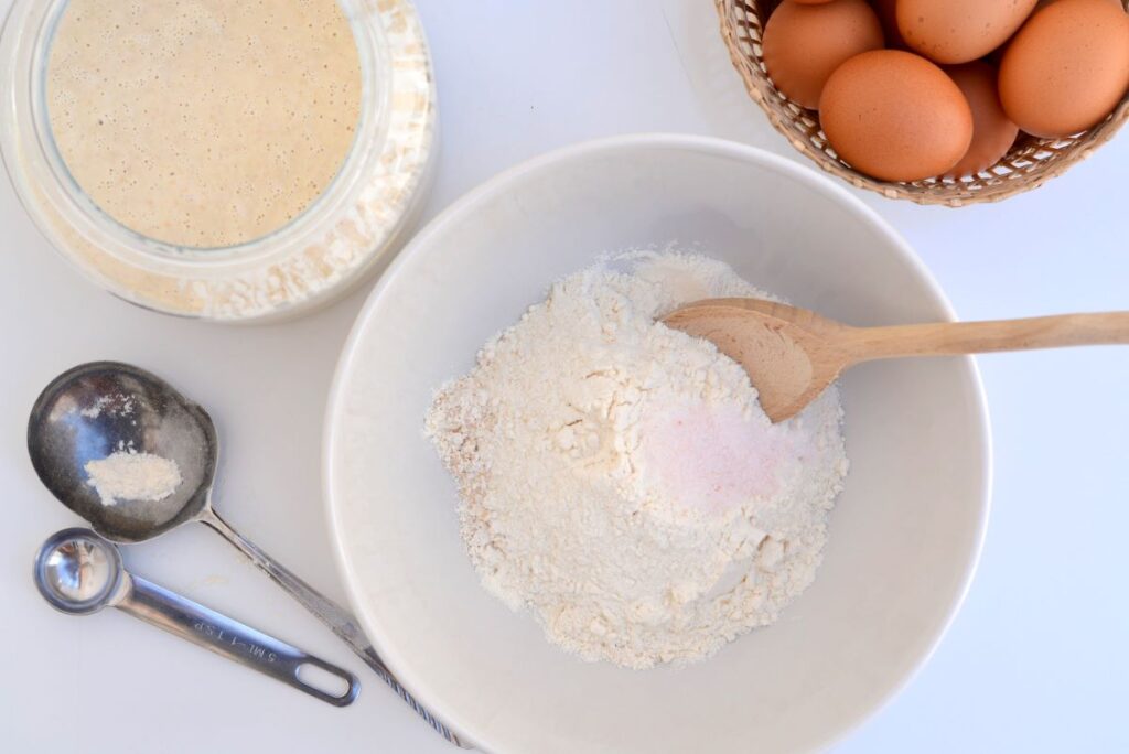 Witte kom met bloem en zout staat op tafel naast een glazen pot met zuurdesem en een mandje met eieren.