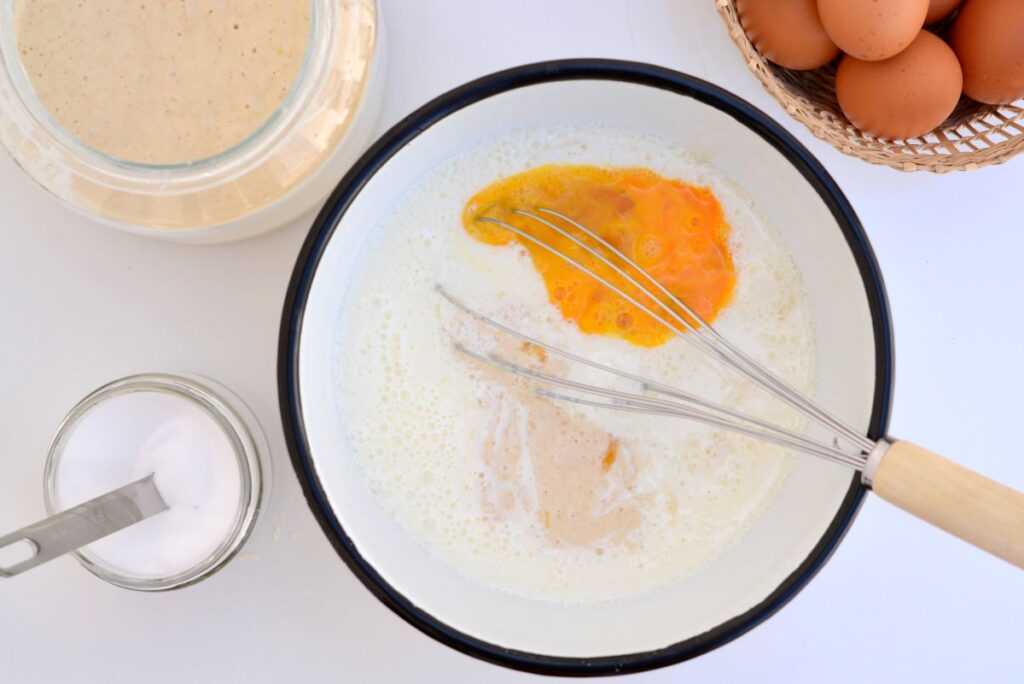 Kom met beslag van karnemelk, zuurdesem, ei en honing dat nog gemixt moet worden met een garde. De kom staat op tafel naast een glazen pot met zuurdesem en een mandje met eieren.