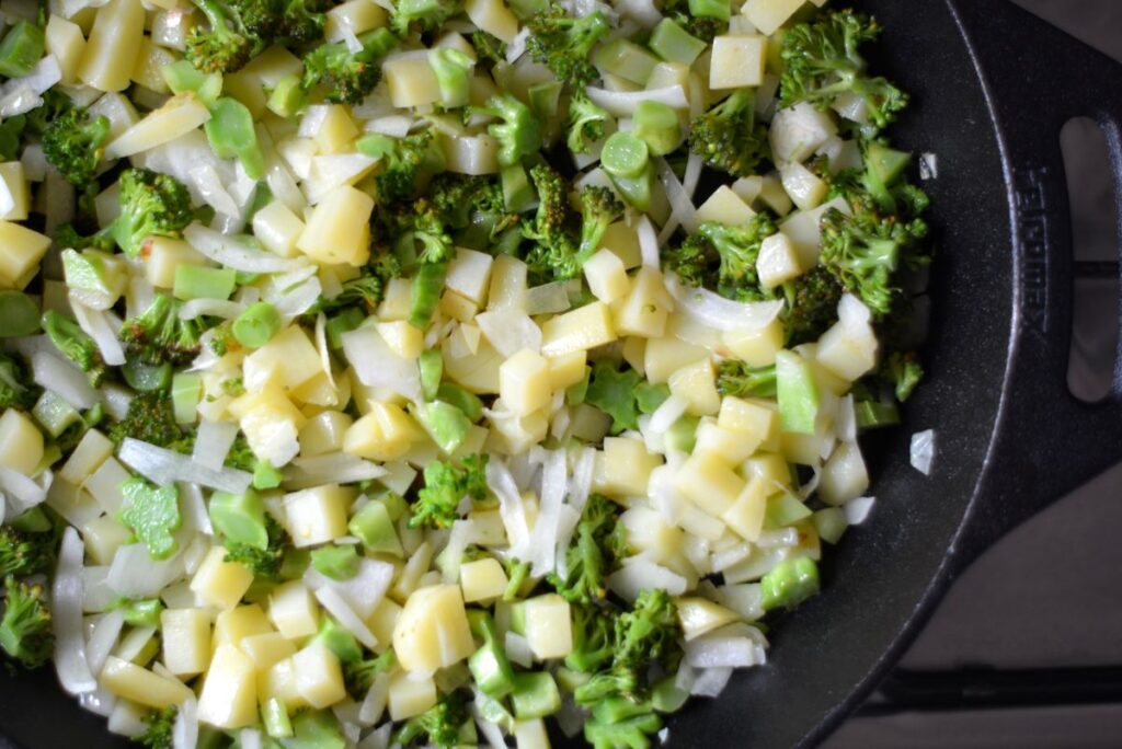 stukjes broccoli, aardappel en ui worden gebakken in een gietijzeren pan 