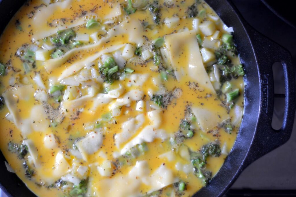 stukjes broccoli, aardappel en ui worden in gietijzeren pan bedekt met plakjes kaas en geklutst ei voor frittata oven recept