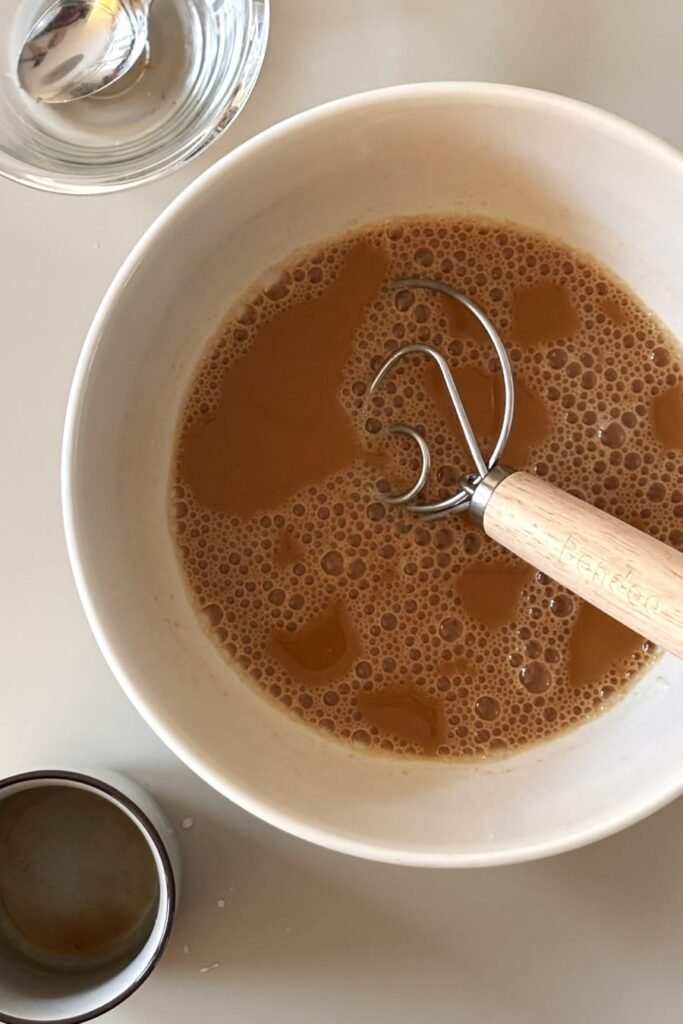 Een mengsel van warm water, koffie, suiker en zuurdesemstarter ligt in een witte schaal op tafel. 