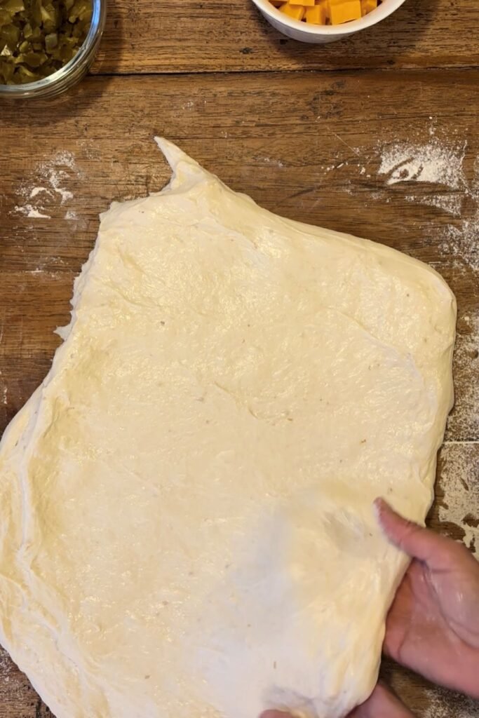 Gerezen tarwebloem deeg wordt uitgespreid over een houten tafelblad. Een potje met cheddar kaas en een potje met gesneden jalapeño pepers staan klaar.