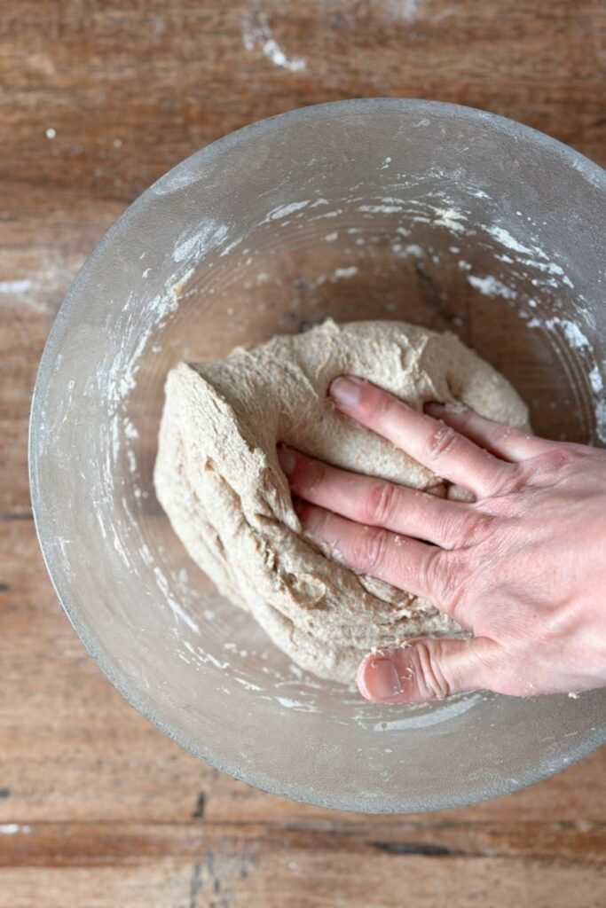 Vrouw gaat zuurdesem brood maken en vouwt met haar hand het deeg voor volkoren brood in een glazen schaal