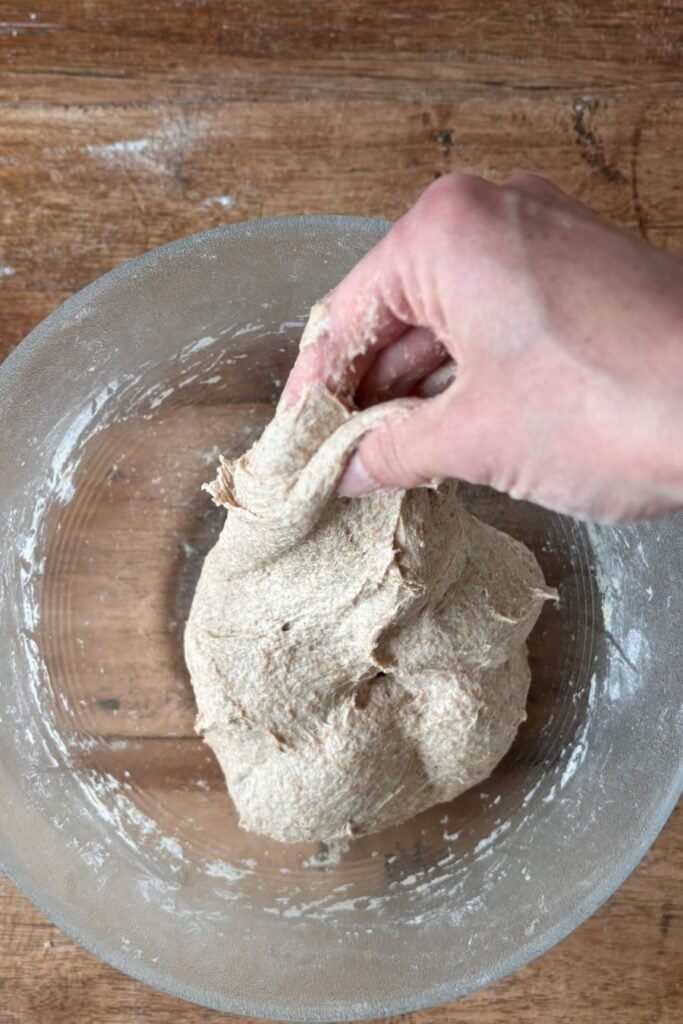 Vrouw gaat zuurdesem brood maken en vouwt met haar hand het deeg voor volkoren zuurdesem brood in een glazen schaal