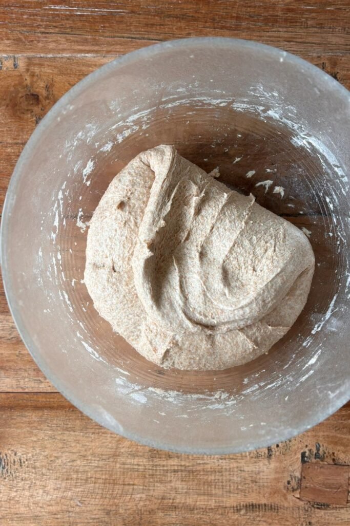 Gevouwen volkoren deeg voor zuurdesem brood ligt in een glazen schaal op tafelblad.