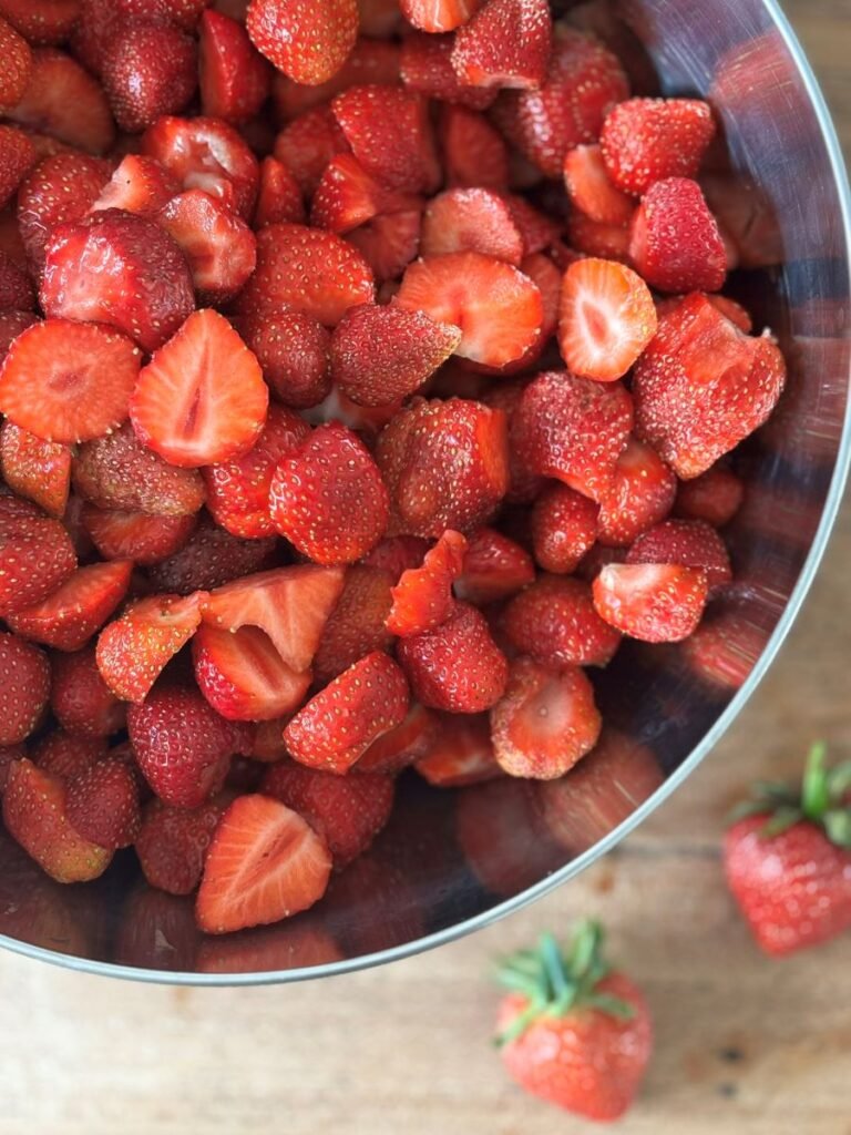 Gesneden aardbeien liggen in een grote bak op een houten eettafel klaar om aardbeienjam van te maken