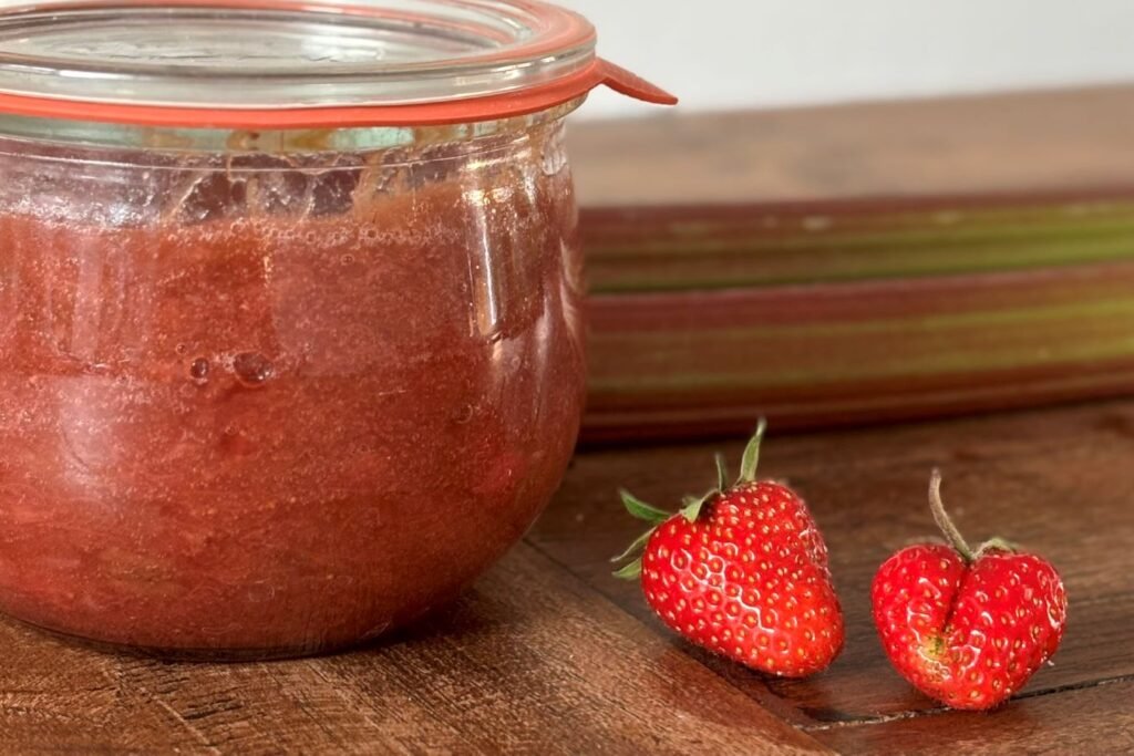 Weckpot met rabarberjam met aardbeien staat op houten tafel naast 2 stengels rabarber en 2 aardbeien.