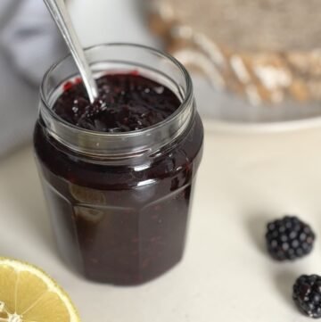 Glazen pot met eigen gemaakte bramenjam staat op een wit tafelblad naast 3 verse bramen, een doorgesneden citroen en 2 zuurdesembroodjes op een wit bord