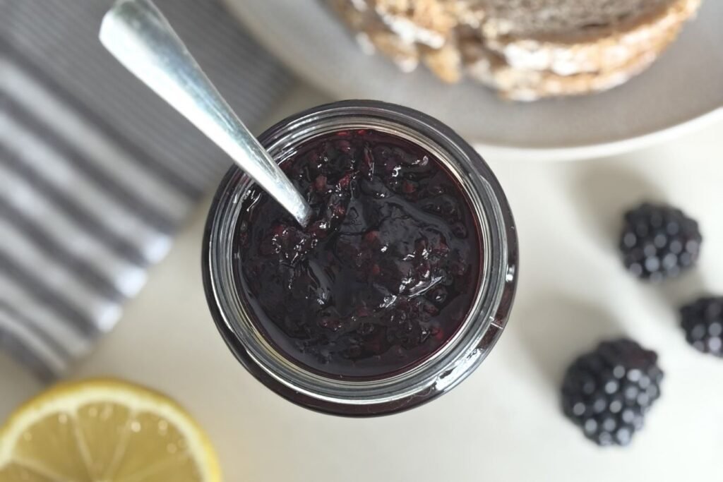 Glazen pot met zelfgemaakte bramenjam en opstaande lepel staat op een wit tafelblad naast een gesneden citroen, 3 verse bramen en 2 zuurdesembroodjes op een bord. 