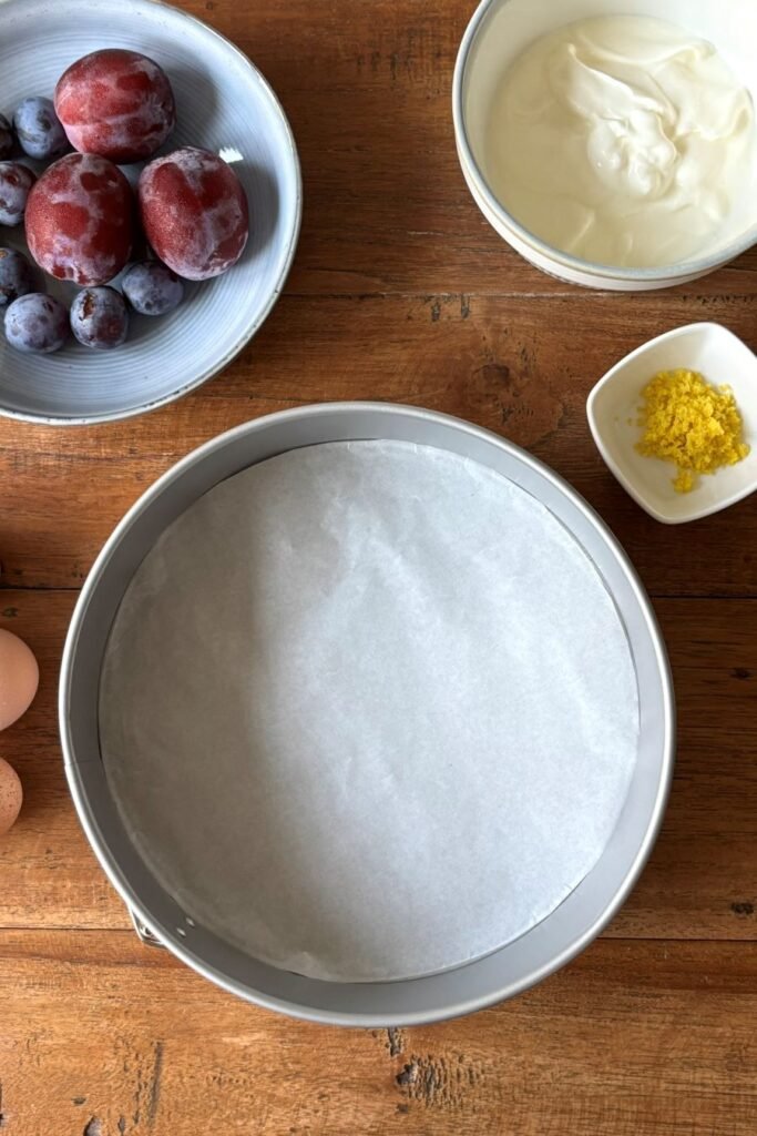 Springvorm staat op tafel klaar met pruimen, yoghurt en citroenrasp voor het bakken van een pruimen cake