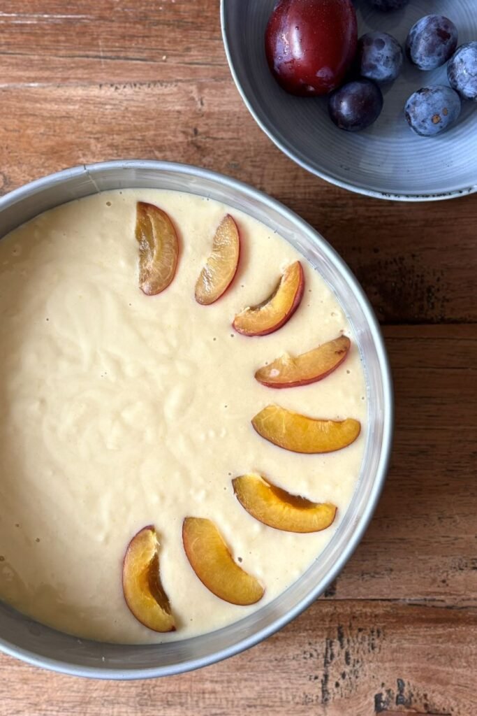 Springvorm gevuld met beslag voor pruimen cake en schijfjes pruimen staat op houten eettafel. 