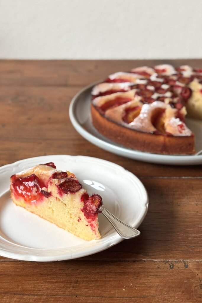 Gesneden stukje pruimen cake ligt op gebaksschoteltje klaar om opgegeten te worden. Op de achtergrond staat de volledige pruimen cake op een groot bord