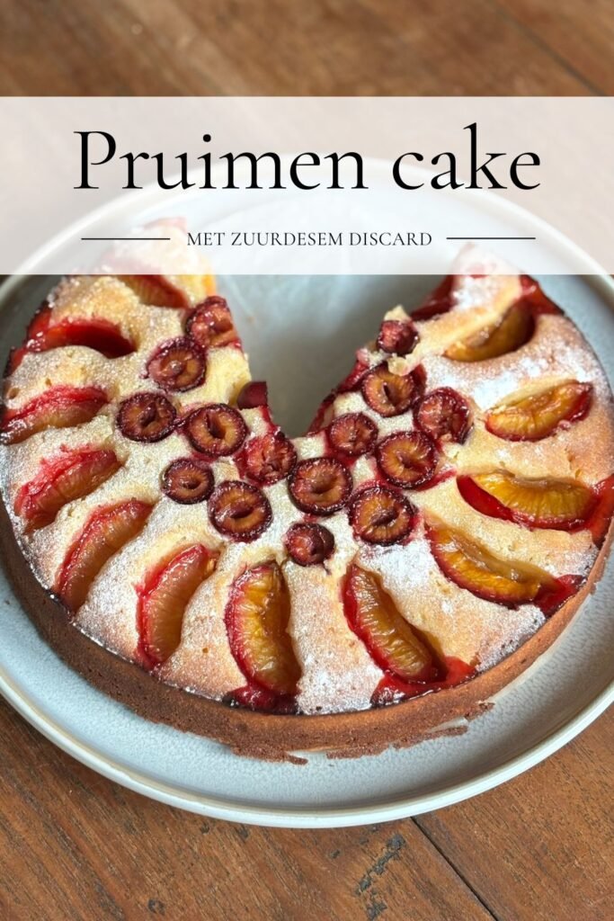 Versgebakken ronde pruimencake bestrooid met poedersuiker ligt op een grijsblauw bord op een houten eettafel