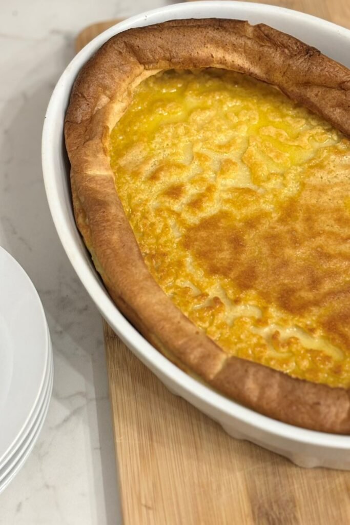 Versgebakken Dutch Baby met zuurdesem in witte ovale ovenschaal staat op een houten plank op een wit keukenblad naast een stabel gebaksbordjes. 