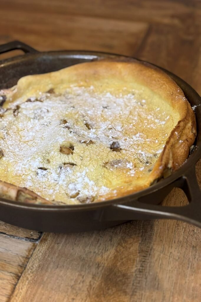 Versgebakken Dutch Baby pannenkoek met zuurdesem bestrooid met poedersuiker ligt in een gietijzeren pan op een houten tafelblad. 