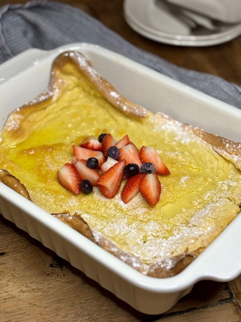 Versgebakken Dutch Baby pannenkoek met zuurdesemdiscard ligt in een witte ovenschaal bestrooid met poedersuiker en gedecoreerd met stukjes aardbei en blauwe bessen.