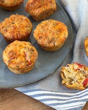 Hartige muffins gevuld met geroosterde paprika, feta, geraspte kaas, olijven en lente uit liggen op een blauw serveer bord naast een blauw gestreepte theedoek