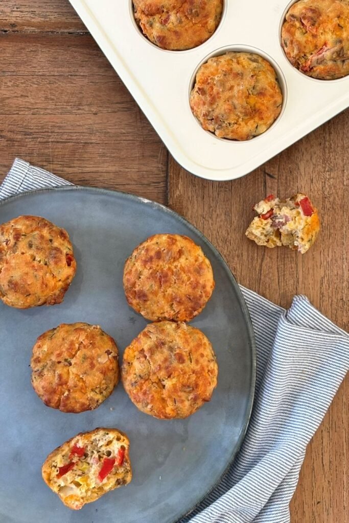 Hartige taartjes in muffinvorm liggen op een houten tafelblad af te koelen naast een bord waarop al een aantal hartige muffins met spek en groente liggen